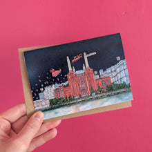 Load image into Gallery viewer, A hand holding a Christmas card in front of a dark pink background. The card is of Battersea Power Station with Santa flying overhead.