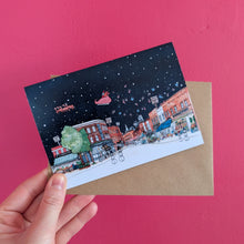 Load image into Gallery viewer, A hand holds a sChristmas Card of Broadway Market in Hackney, London, featuring a snowy night sky with Santa and his sleighs, layered over a brown kraft envelope against a bright pink background.