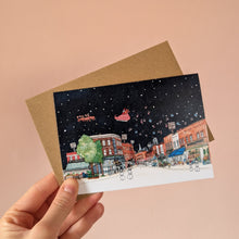 Load image into Gallery viewer, A snowy Broadway Market scene on a Christmas card showing one of Hackney's most loved markets with Santa flying overhead. The card is held with a brown kraft envelope in front of a light pinky peach wall.
