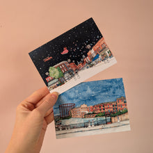 Load image into Gallery viewer, A hand holds two illustrated art cards against a plain peach-pink wall. One card shows a snowy night city scene of Broadway Market with a red Santa sleigh against a dark sky, and the other shows a snowy day city scene of Regent's Canal.