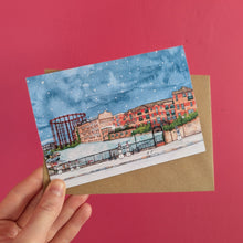 Load image into Gallery viewer, A hand holding a watercolour Christmas card of Regents Canal in Hackney with the old gas works against a snowy blue sky.