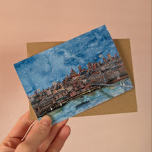 Load image into Gallery viewer, A hand holds an A6 watercolour art Christmas card featuring a snowy city waterfront scene in blue tones, layered over a brown kraft envelope against a soft peach-pink background.