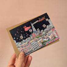 Load image into Gallery viewer, A hand holds an A6 Christmas card featuring a whimsical London cityscape at night with a red sleigh carrying Santa Claus past The Shard, shown against a plain warm peach background with a brown kraft envelope.