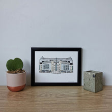 Load image into Gallery viewer, A framed print of Chelsea FC football ground. A detailed fineliner drawing and watercolour painting. In a black frame, on a wooden desk with a small plant next to it. 
