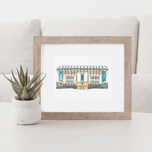 Load image into Gallery viewer, A print of Leeds United football stadium in a wooden frame with a white mount. The print is photographed on a white coffee table. 
