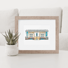 Load image into Gallery viewer, A print of Leeds United football stadium in a wooden frame with a white mount. The print is photographed on a white coffee table.