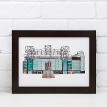 Load image into Gallery viewer, A detailed Old Trafford art print, the piece shows the front of the football stadium home to Manchester United FC. Framed in a black frame and propped against a white brick wall.