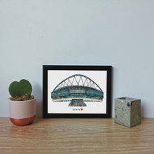 Load image into Gallery viewer, A hand-drawn print of Wembley Stadium, where England's Women's Football team won the 2022 Euros. The piece has the text 'It's Home' with a red heart underneath it. It is in a black frame on a desk.