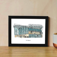 Load image into Gallery viewer, A full colour print of Newcastle United FC, the print shows St James' Park football ground with 'The Magpies' underneath. It is in a black frame on a wooden table.