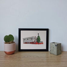Load image into Gallery viewer, A hand drawn and painted print of Nottingham Forest Football Ground. The illustration is framed in a blacck frame, photographed on a desk next to a small pot plant.