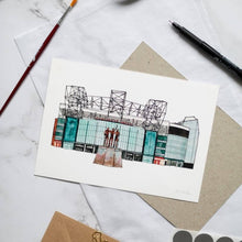 Load image into Gallery viewer, An Old Trafford print, the piece is a hand painted illustration of the football stadium by Jessica Sian. It is photographed on a desk with a pen and paintbrush beside it.

