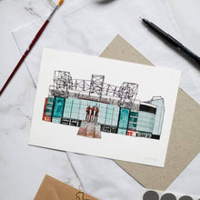 Load image into Gallery viewer, An Old Trafford print, the piece is a hand painted illustration of the football stadium by Jessica Sian. It is photographed on a desk with a pen and paintbrush beside it.