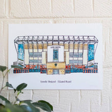 Load image into Gallery viewer, A personalised print of Elland Road, this piece of Leeds United wall art has been personalised with the club name and ground typed beneath the painting. Photographed on a white brick wall with a pot plant showing in the corner.
