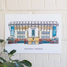Load image into Gallery viewer, A personalised print of Elland Road, this piece of Leeds United wall art has been personalised with the club name and ground typed beneath the painting. Photographed on a white brick wall with a pot plant showing in the corner.