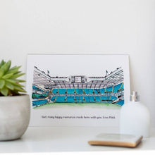 Load image into Gallery viewer, A detailed pen and watercolour illustration of White Hart Lane, Tottenham Hotspur's old football ground. The print is personalised with a personal message and is on a white dressing table beside a small plant, phone and bottle of perfume.