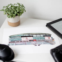Load image into Gallery viewer, An Arsenal football stadium print on a bedside table. The piece is surrounded by a plant, kindle, headphones and games console controller.