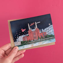 Load image into Gallery viewer, A hand holding a Christmas card featuring Battersea Power Station under a dark night sky with santa flying overhead, set against a solid bright pink background.
