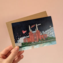 Load image into Gallery viewer, A Christmas card of Battersea Power Station by Jessica Sian Illustration. The card is held against a brown raft envelope with a light pink wall behind it. 

