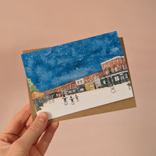 Load image into Gallery viewer, London's famous Columbia Road is shown in the Christmas card here, held against a brown kraft envelope, against a light pink wall. The card is Christmas themed with snow and snowmen in it. 
