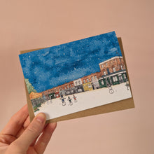 Load image into Gallery viewer, A hand holding an A6 Christmas Card with a watercolour print of a snowy Columbia Road with snowmen under a snowy blue sky, set against a solid peach background with a brown envelope.
