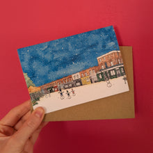 Load image into Gallery viewer, A hand holds a Christmas card of Columbia Road by artist Jessica Sian. The watercolour design shows snowmen in the street and is held with a brown kraft envelope against a dark pink background.
