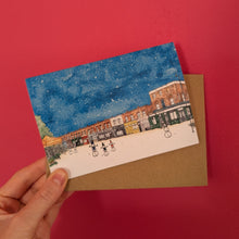 Load image into Gallery viewer, A hand holds an A6 Christmas card of a watercolour scene of Columbia Road Flower Market in London. Snowmen play in the scene, under the blue snowy sky. The card is shown with a brown kraft envelope behind it against a solid red background.
