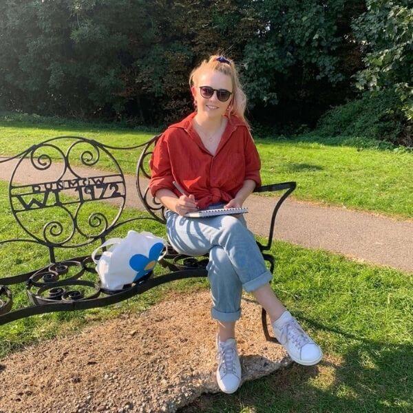 Jessica Sian, the artist behind Jessica Sian Illustration, sitting on a bench drawing in Dunmow. It's a sunny day.