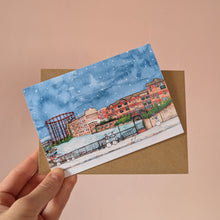 Load image into Gallery viewer, A hand holds a watercolour Christmas card of Regent's Canal in Hackney. It's a snowy scene with snowmen, held with a brown kraft envelope in front of a  peach background.
