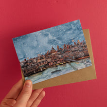 Load image into Gallery viewer, A hand holds a Christmas card featuring a snowy London cityscape over water, looking out to St Paul's Cathedral, with a brown envelope behind it, against a solid pink background.
