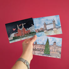 Load image into Gallery viewer, A hand holding three illustrated Christmas Cards showing London landmarks against a solid red background. Featuring Battersea Power Station, Tower Bridge and Trafalgar Square.
