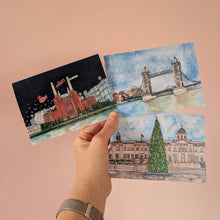 Load image into Gallery viewer, A hand holding three small illustrated Christmas cards against a solid peach-pink background. The postcards depict London landmarks: Battersea Power Station at night, Tower Bridge in the snow, and Trafalgar Square with the Christmas tree.
