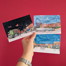 Load image into Gallery viewer, A hand holds three watercolor Christmas cards depicting snowy London market street scenes at night, against a solid red background.
