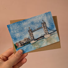 Load image into Gallery viewer, A hand holds a small watercolor-style card featuring Tower Bridge in London, with a brown envelope behind it, against a soft peach-pink background.
