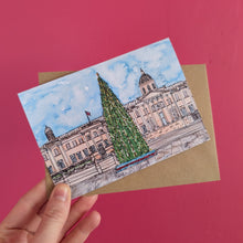 Load image into Gallery viewer, A hand holds a Christmas card featuring a watercolour illustration of Trafalgar Square and its Christmas tree against a solid pink background.
