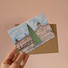 Load image into Gallery viewer, A hand holds an A6 Christmas card showing the National Gallery in Trafalgar Square with a Christmas tree in front. The card is layered over a brown envelope against a soft peach-pink background.
