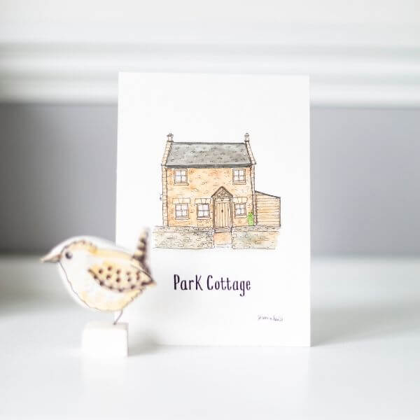 A fineliner and watercolour house painting of a cottage with Park Cottage written underneath. On a white table against a grey wall with a little bird ornament.