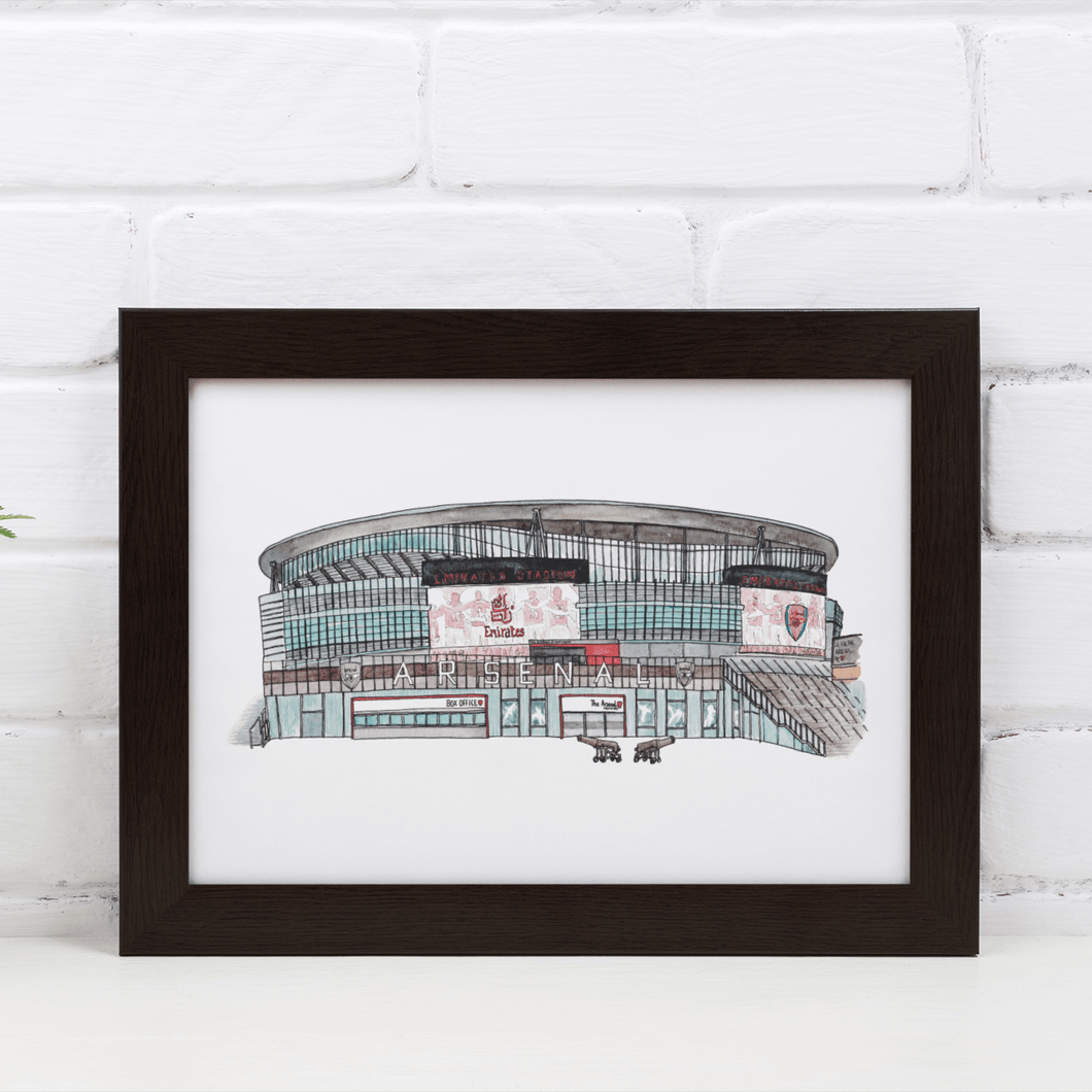 A framed print of the Emirates Stadium, showing the front of the Arsenal FC ground. The print is in a white frame against a white brick wall, on a white table.
