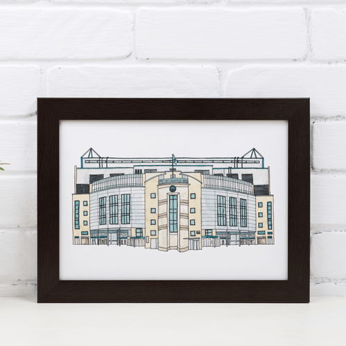 A framed A4 Chelsea Football print, a detailed illustration of the stadium. Framed in a black frame, the frame is propped against a white brick wall.