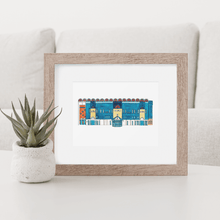 Load image into Gallery viewer, A print of Maine Road football stadium in a wooden frame. The piece is sat on a white coffee table with a small plant beside it.

