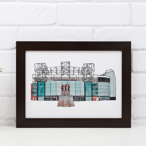 A detailed Old Trafford art print, the piece shows the front of the football stadium home to Manchester United FC. Framed in a black frame and propped against a white brick wall.