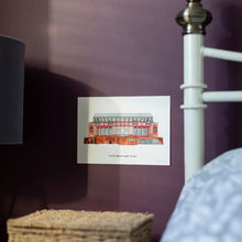 Load image into Gallery viewer, A personalised print of Liverpool Football Stadium with the wording &#39;You&#39;ll Never Walk Alone&#39; written underneath it. The A4 print is displayed on a deep purple wall in a bedroom. 
