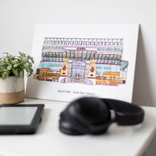 Load image into Gallery viewer, West Ham United wall art by Jessica Sian, a detailed illustration of the Upton Park football stadium. The piece is on a bedside table with headphones, a kindle and a small pot plant.
