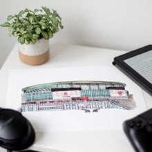 Load image into Gallery viewer, An Arsenal football stadium print on a bedside table. The piece is surrounded by a plant, kindle, headphones and games console controller.
